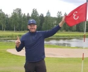 Mand på mørk trøje og kasket holder et flag ved golfhul 3 og viser en finger, med golfbane og træer i baggrunden.