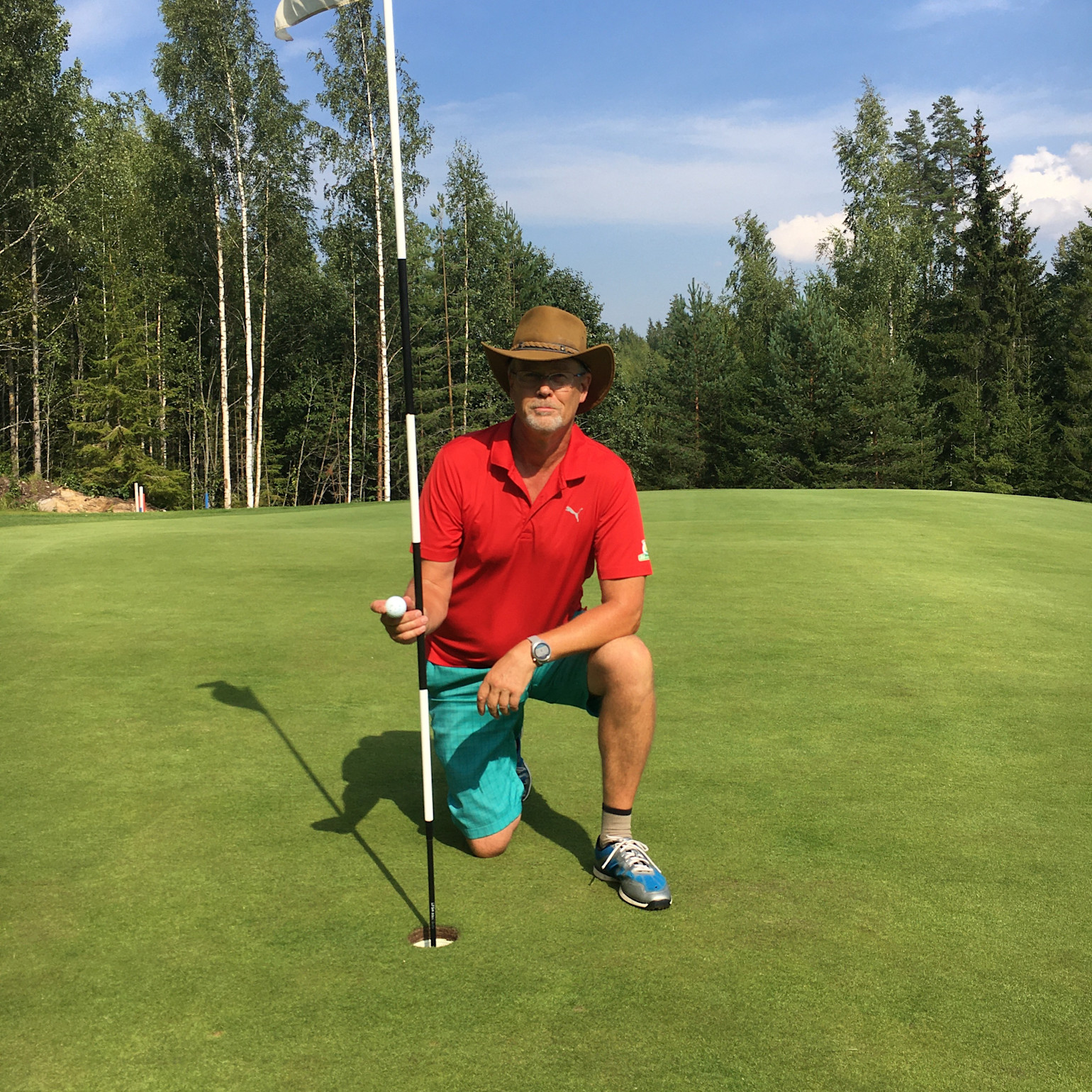 Mand i rød poloshirt og hat knæler ved et golfhul, holder en golfbold og flagstang på en grøn golfbane omgivet af skov.