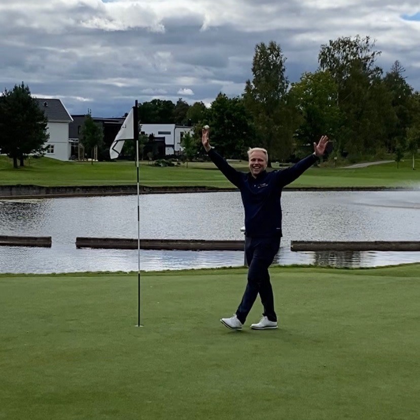 Man firar med armarna uppsträckta bredvid en golfflagga på en green, med en damm och träd i bakgrunden.