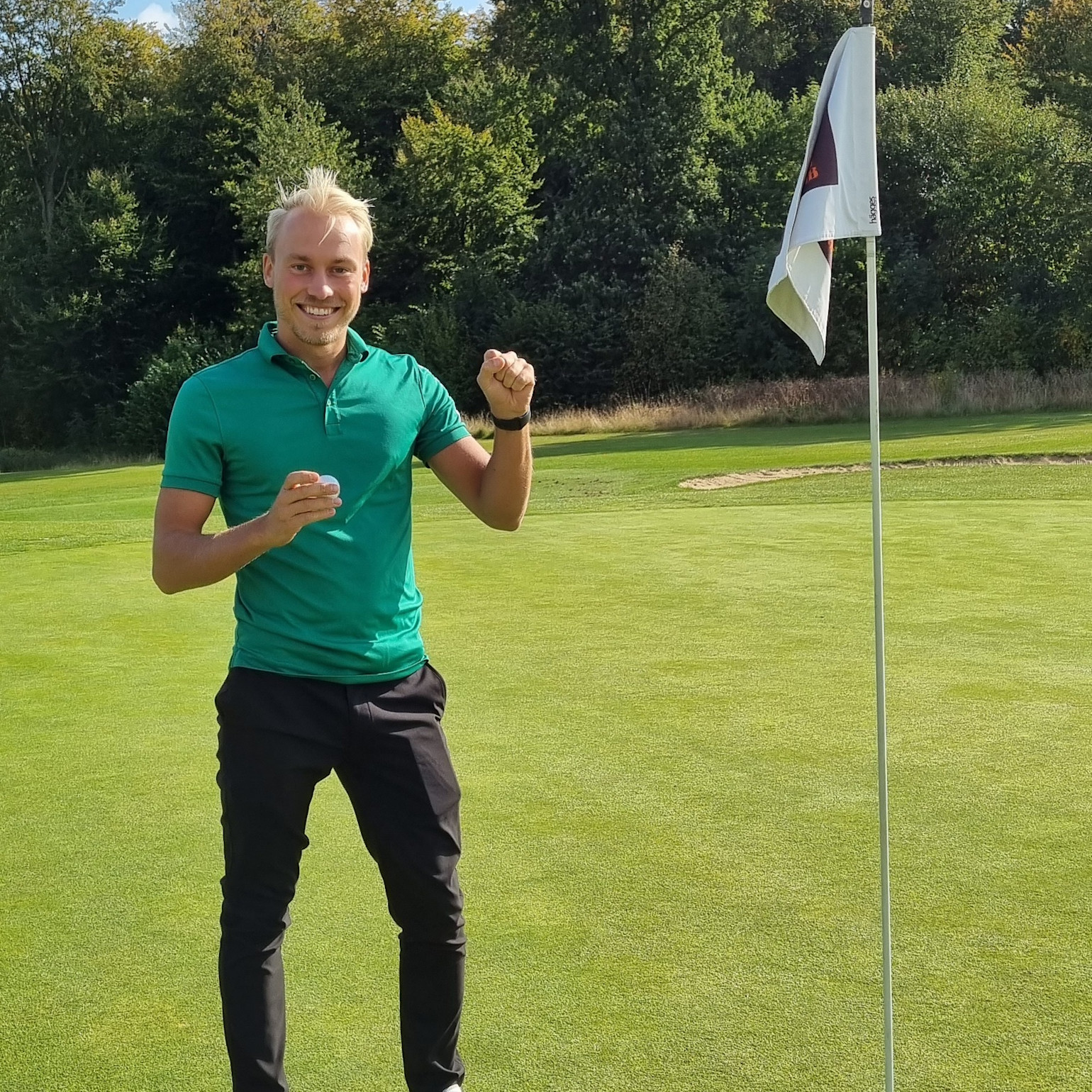 Mand i grøn poloshirt fejrer ved siden af et golfhulflag på en golfbane med træer i baggrunden.