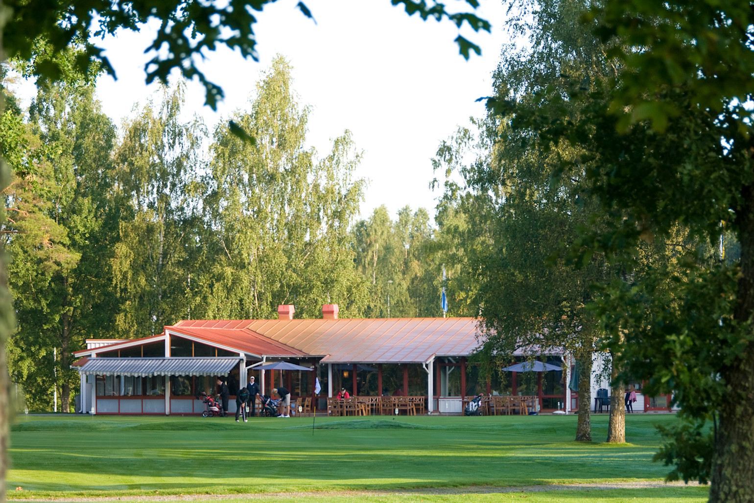 Golfspillere øver på greenen foran en klubhusbygning omgivet af træer og grønne omgivelser på en solrig dag.