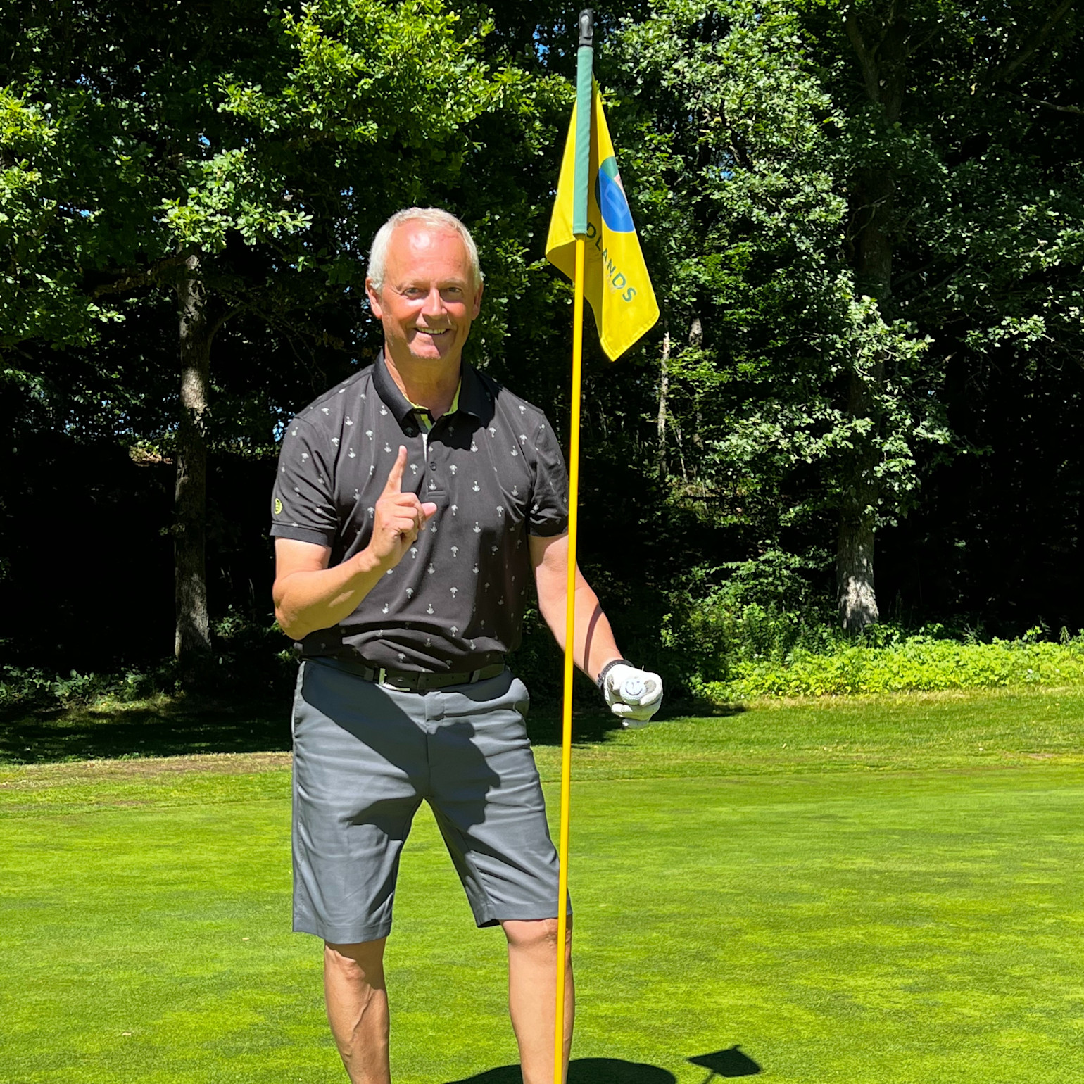 Mand i golfbeklædning står ved siden af en flagstang på en golfbane, holder én finger op og smiler til kameraet.