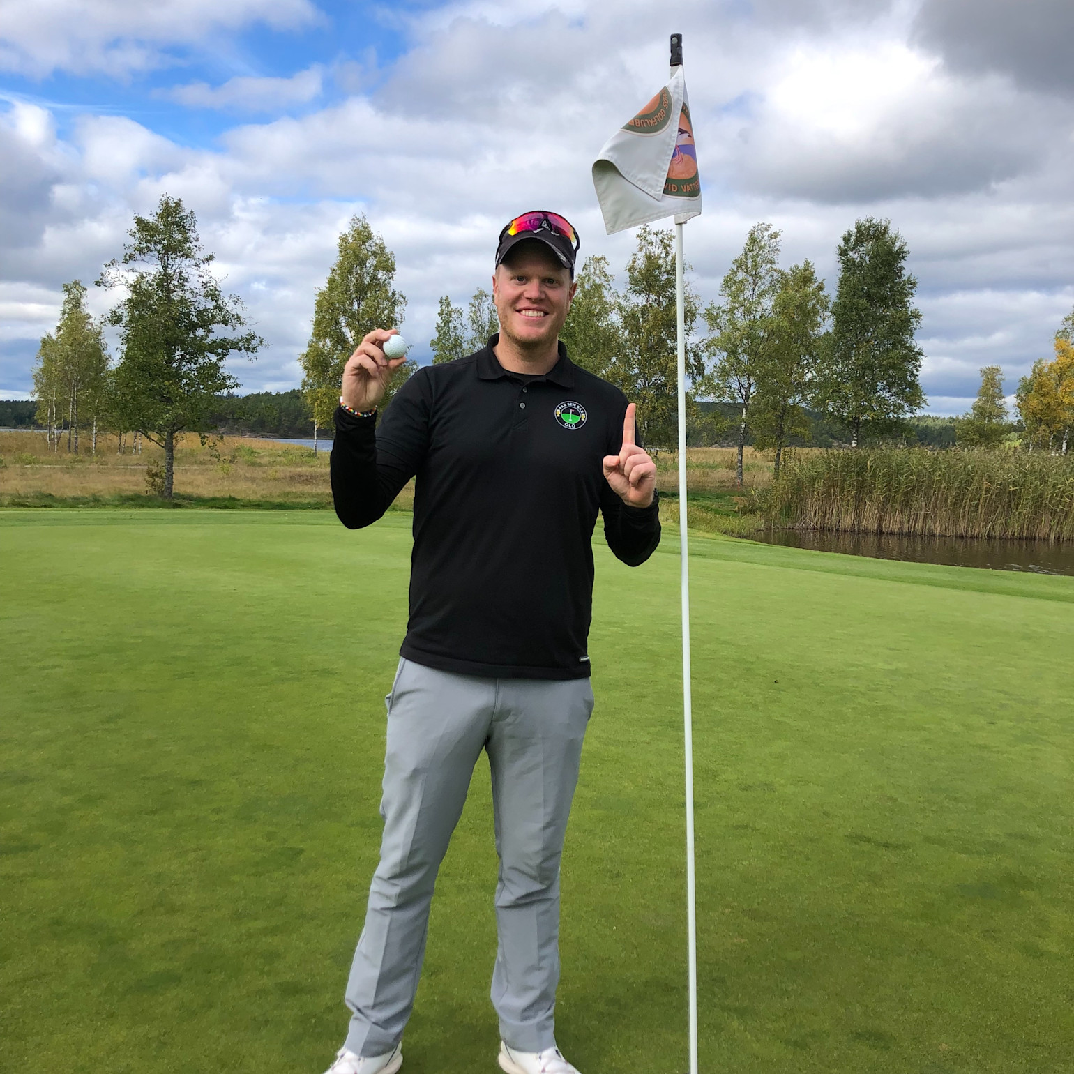 Man i golfkläder håller upp en golfboll och pekar med ett finger bredvid en flaggstång på en golfbana med träd i bakgrunden.
