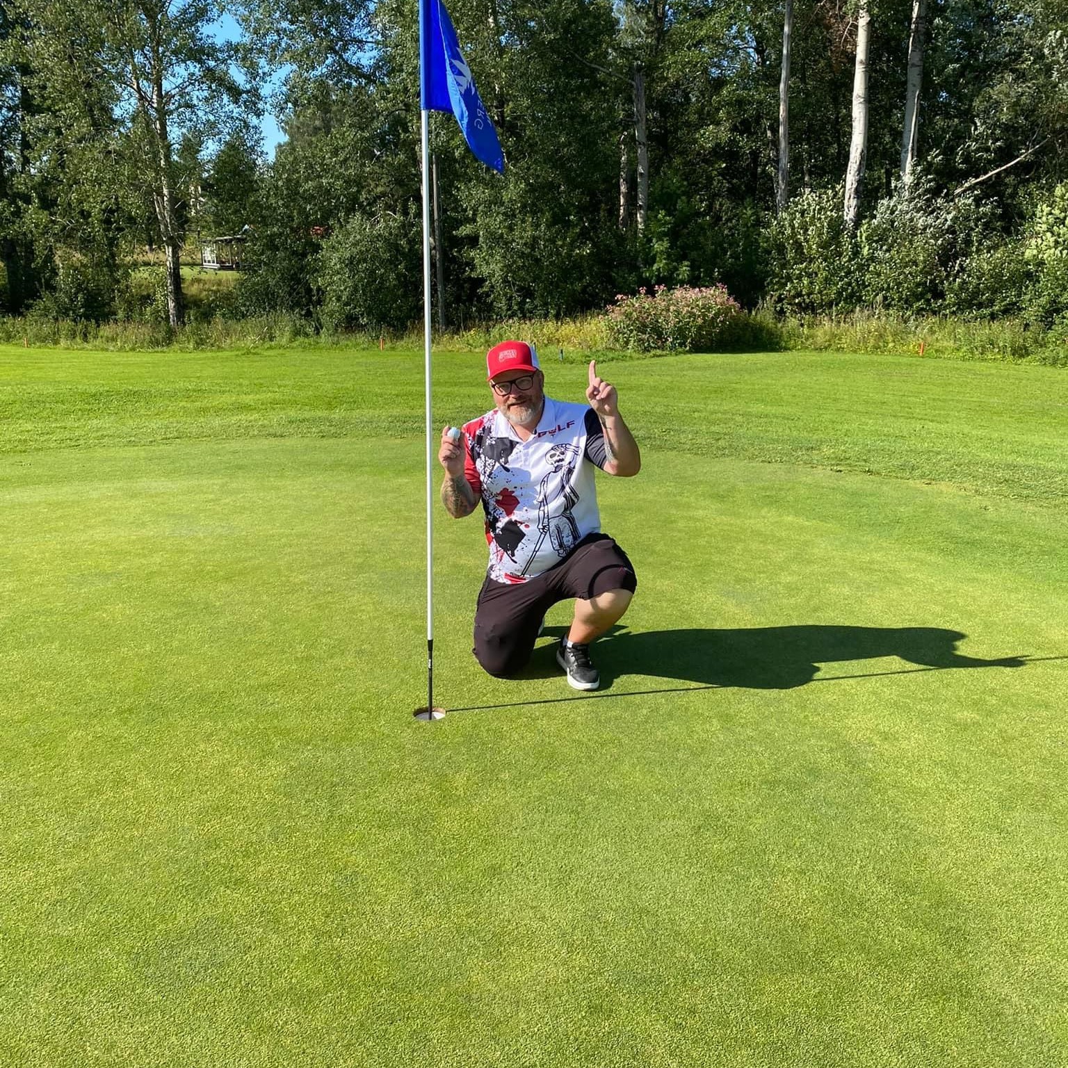 Man i golfkläder knäböjer bredvid ett golfhål med flagga, håller upp en golfboll och pekar uppåt på en grön golfbana.