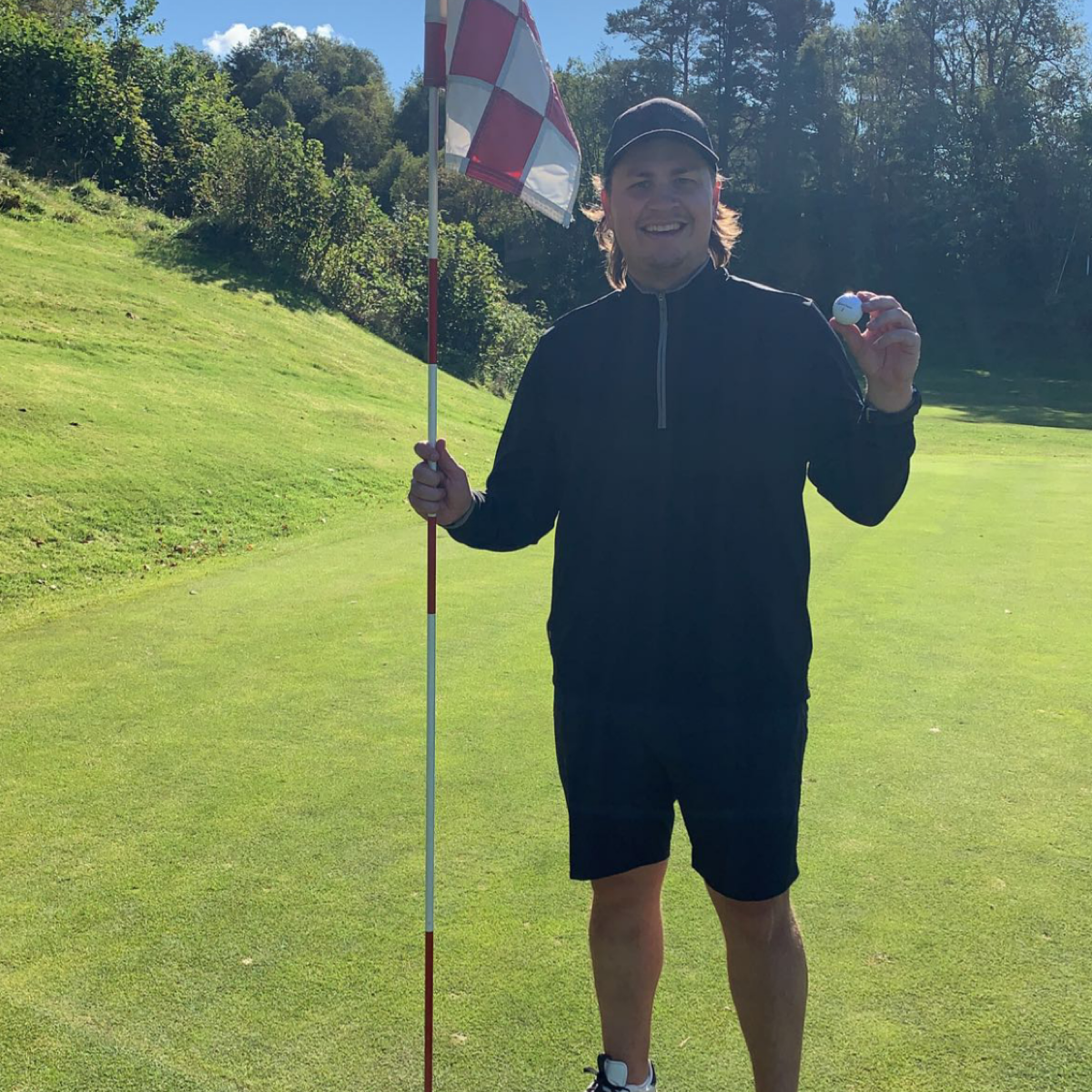 En mand i mørkt tøj holder et golfklubflag i den ene hånd og en golfbold i den anden på en solrig golfbane.