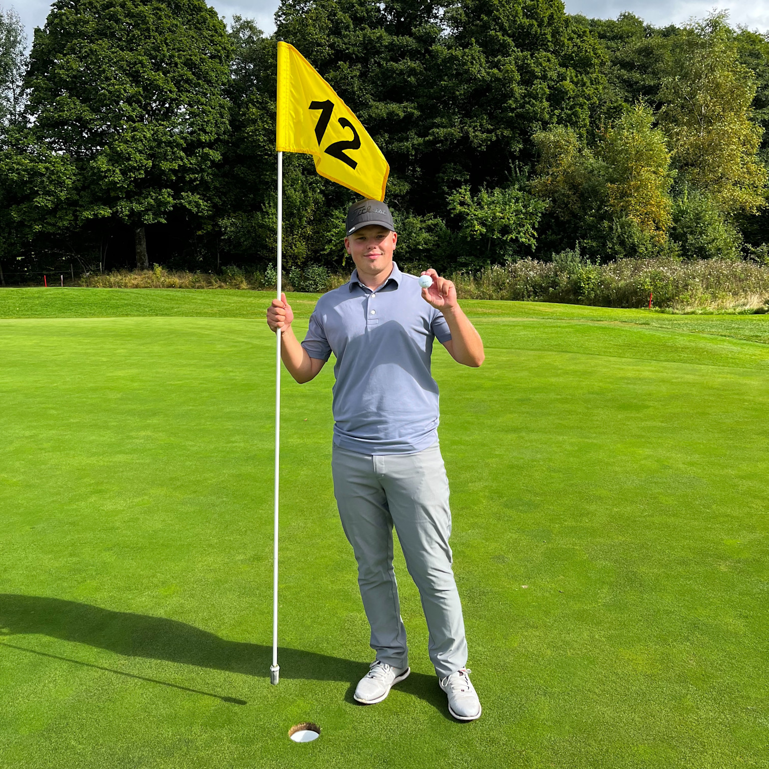 En mann står på en golfbane, holder i flaggstangen med nummer 12 og viser fram en golfball ved siden av hullet.