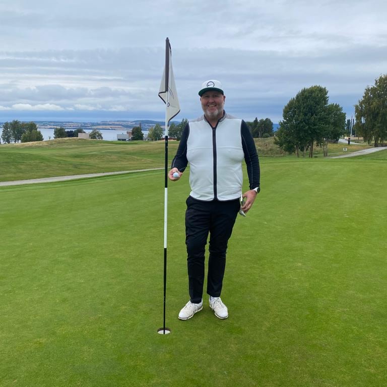 Mand iført golfbeklædning står smilende ved siden af en flagstang på en golfgreen og holder en golfbold, med træer og landskab i baggrunden.