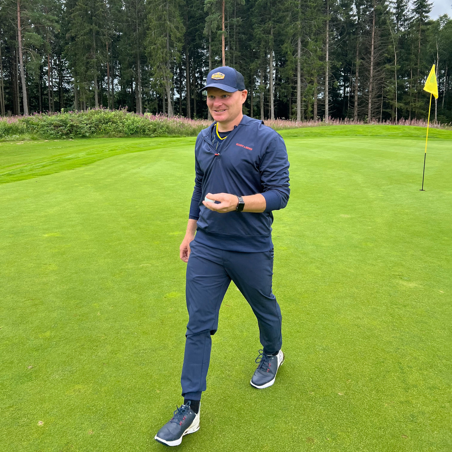 A man in blue golf attire is walking on a golf course with a yellow flag in the background and forest all around.