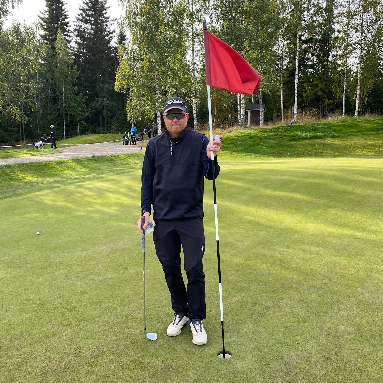 Man holder en golfkølle og en flagstang på en green, omgivet af træer og andre golfspillere i baggrunden.
