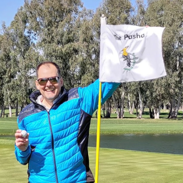 Mand i blå jakke holder en golfbold og en flagstang med et flag, hvor der står "The Pasha", på en golfbane med træer i baggrunden.