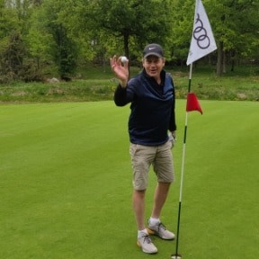 Man holder en golfbold op ved siden af et hul med flag på en golfbane omgivet af grønne omgivelser.