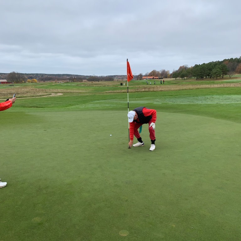 En golfspelare böjer sig ner vid hålet på greenen för att plocka upp sin boll, medan en annan person fotograferar från sidan.