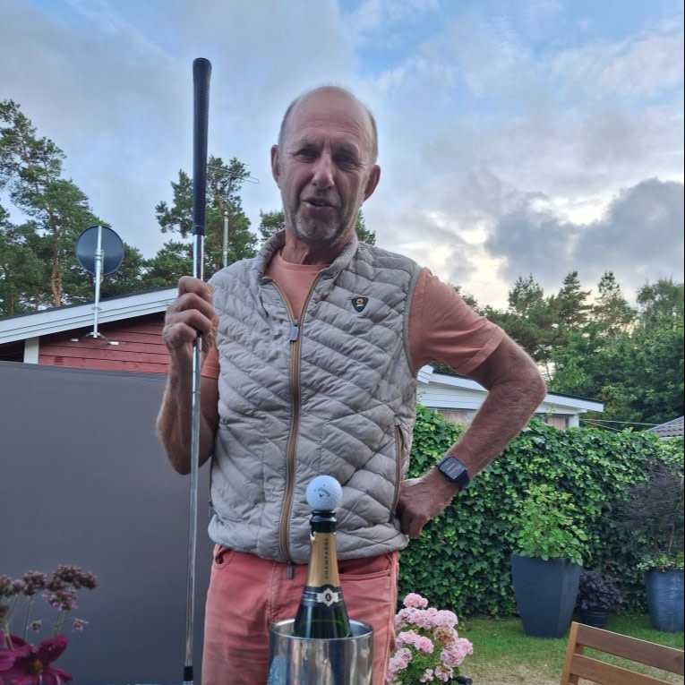 A man with a golf club stands outdoors by a table with a champagne bottle and a golf ball on top, surrounded by plants and flowers.