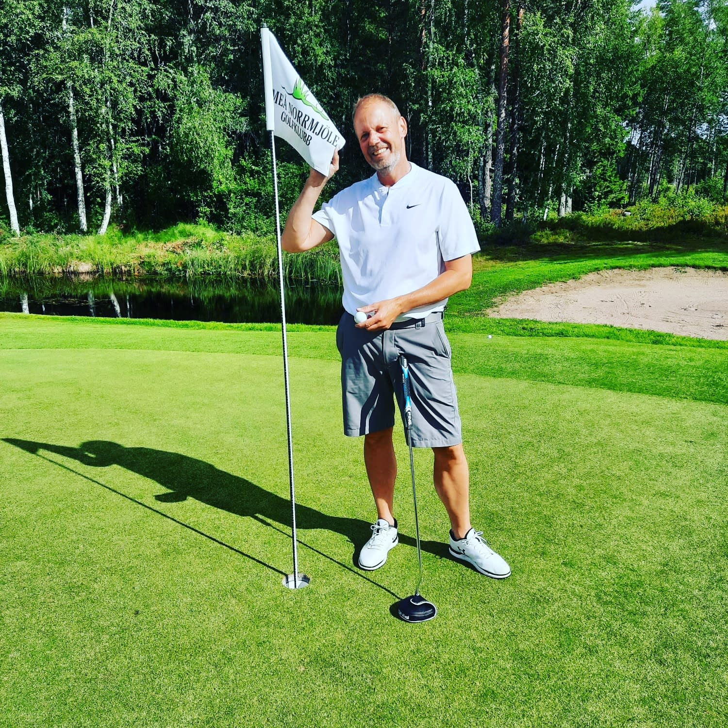 Mand iført en hvid polo og grå shorts holder en golf­flag på greenen, med skov og vand i baggrunden.