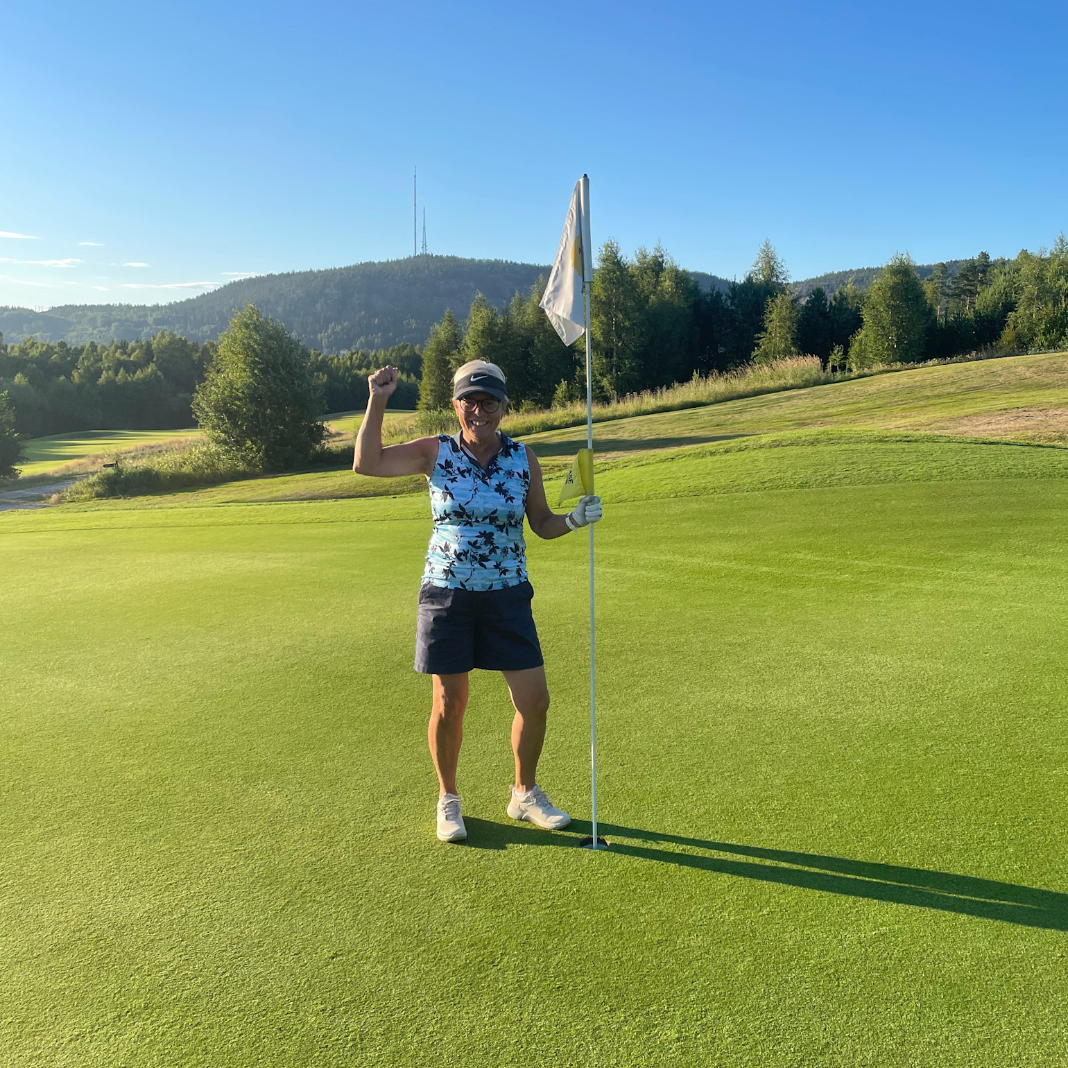 Kvinde i golfbeklædning fejrer ved siden af en flagstang på en golfgreen med skov og bakker i baggrunden.