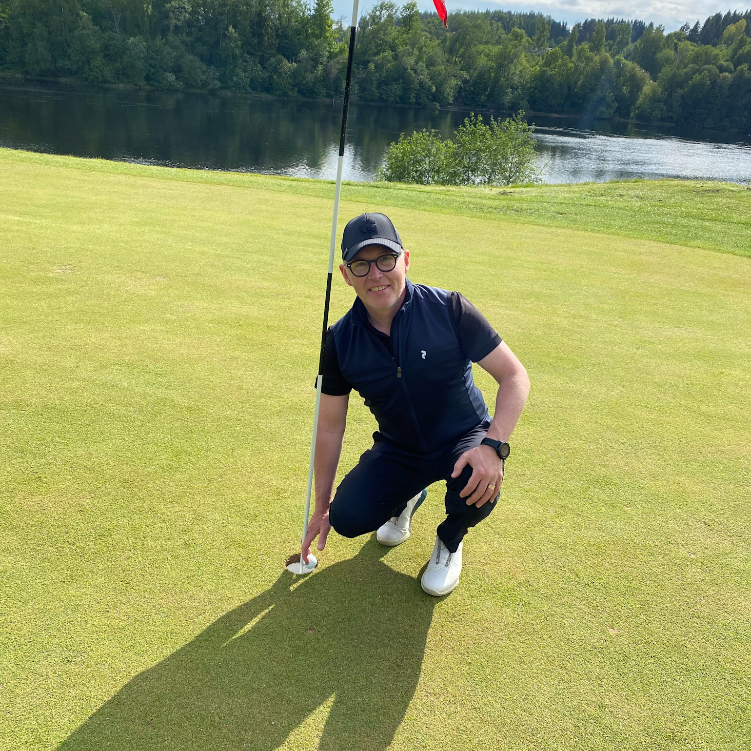 En mand iført golfbeklædning huker sig ned ved et golfhul og holder i flagstangen på greenen, med en sø og skov i baggrunden.
