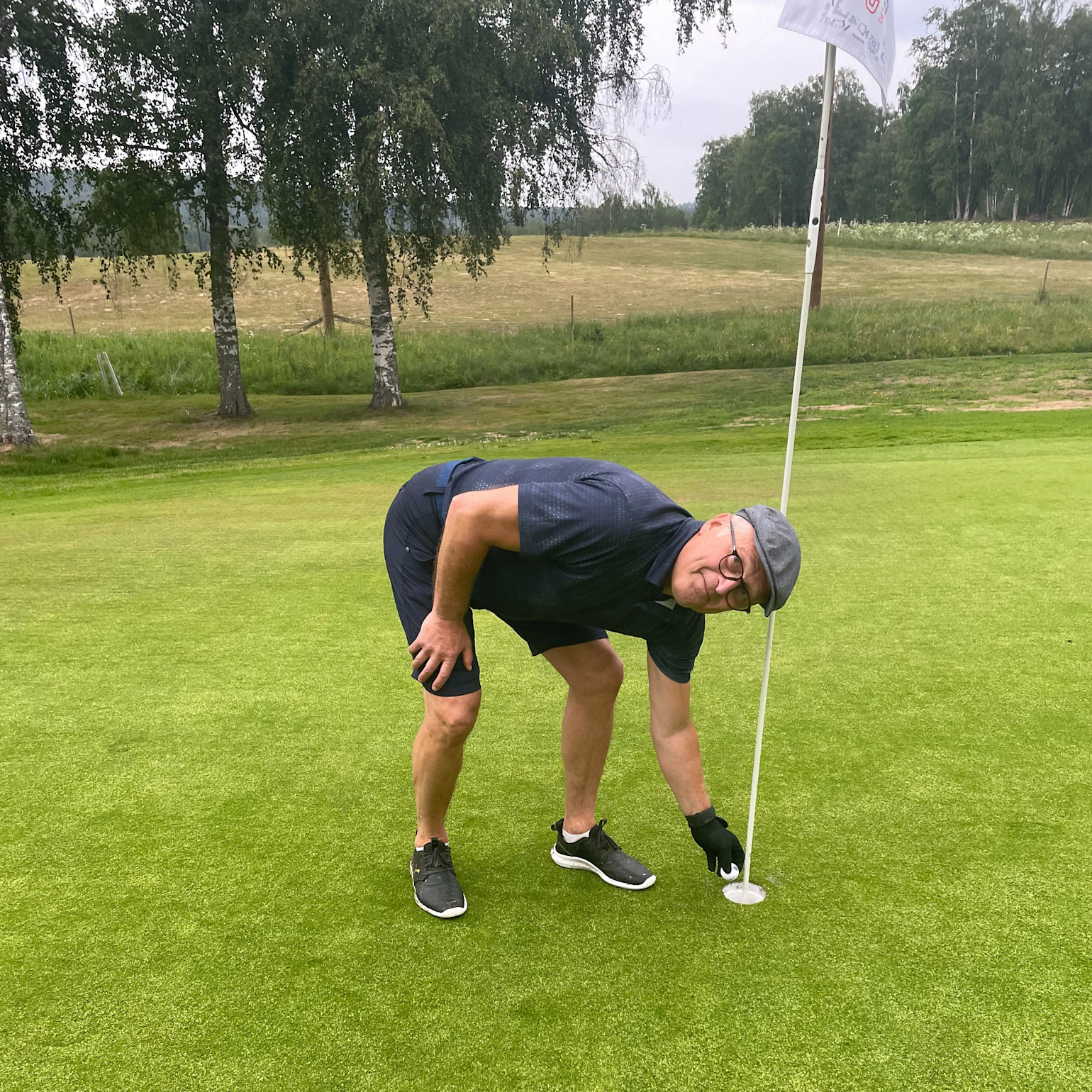 En mand iført golfbeklædning bøjer sig ned og tager en golfbold op af hullet på en green, med flagstang og træer i baggrunden.