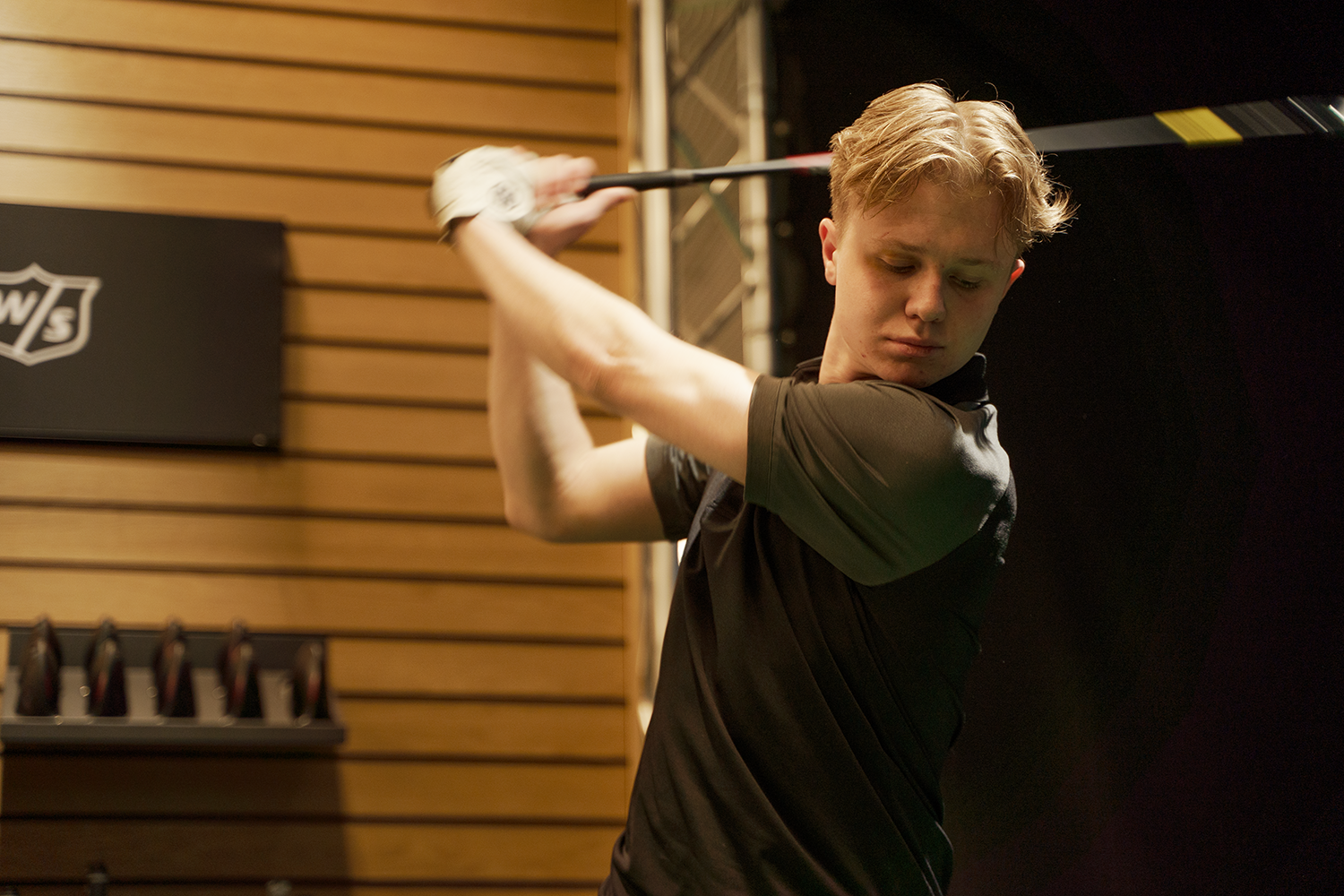 Ung man svingar en golfklubba inomhus, klädd i svart tröja, med träpanelvägg och golfutrustning i bakgrunden.