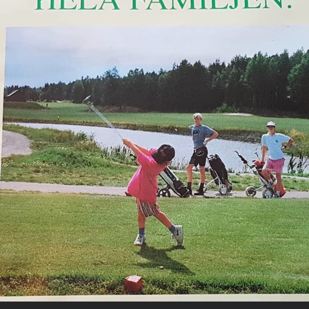 Et barn svinger en golfkølle på en golfbane, mens to voksne med golfvogne ser på ved en sø og en skov.