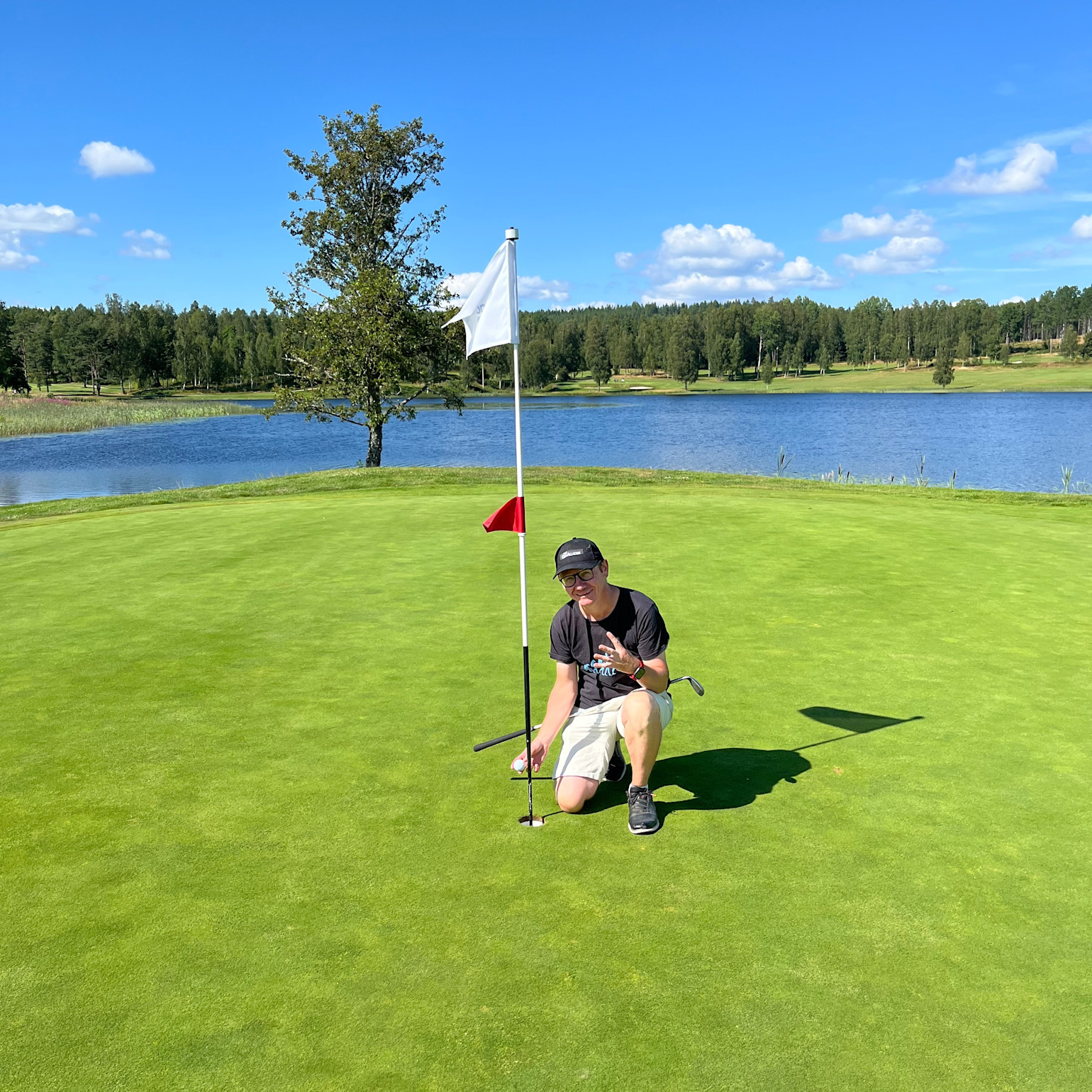 Une personne est accroupie près d’un trou de golf sur le green, tenant un club de golf et une balle, avec un lac et des arbres en arrière-plan sous un ciel bleu.