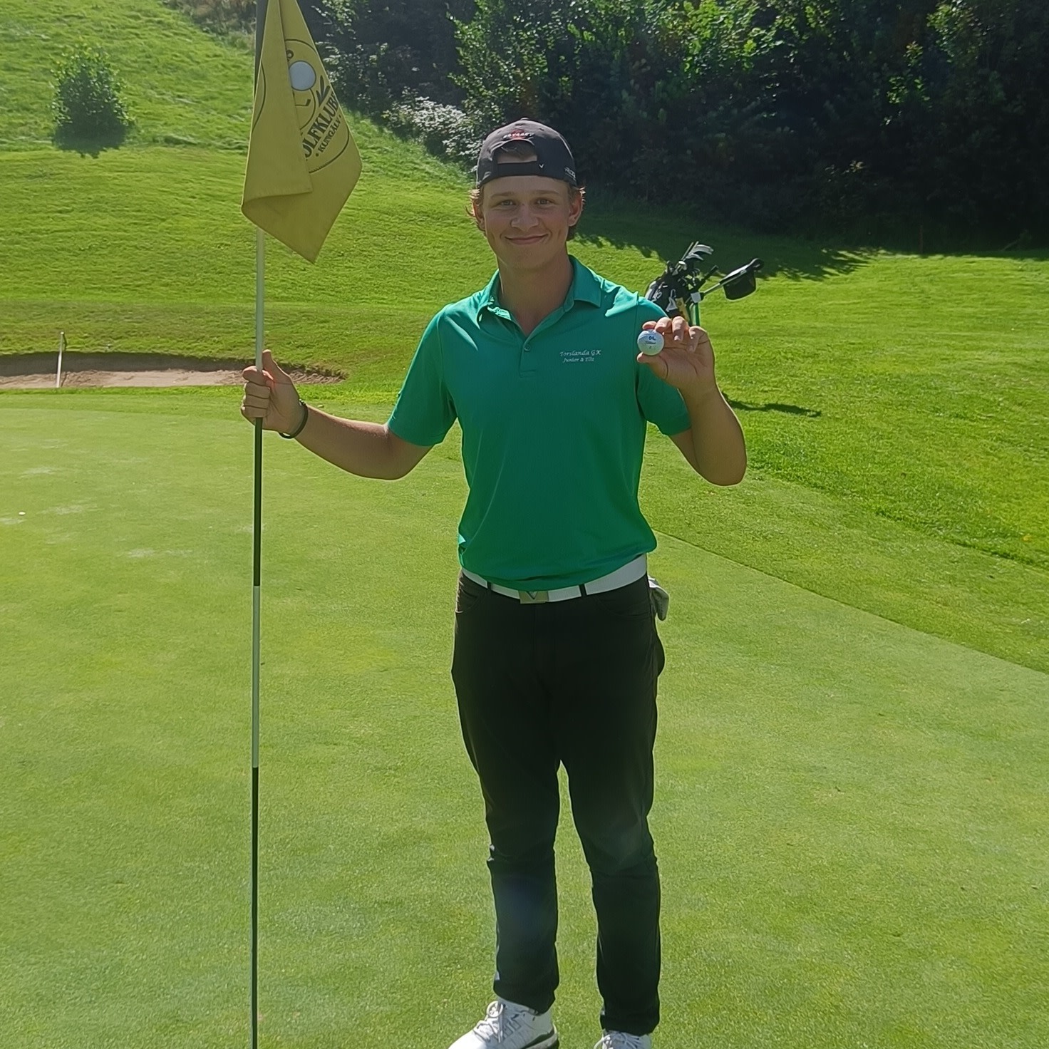 Ung mand i grøn poloshirt og kasket holder en golfbold og flagstang på en golfgreen på en solrig dag.