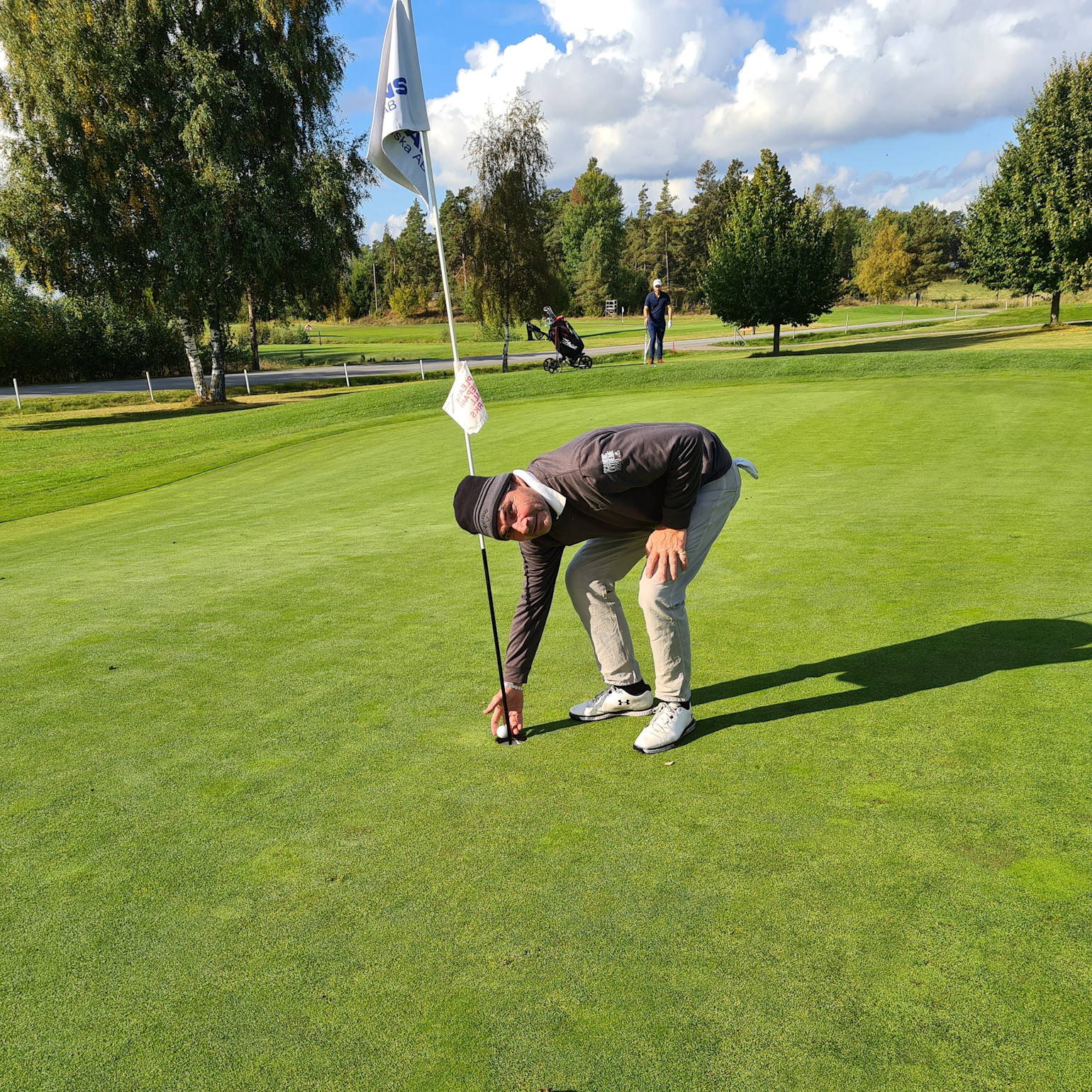 Man böjer sig ner för att plocka upp en golfboll vid hålet på en golfbana under en solig dag.