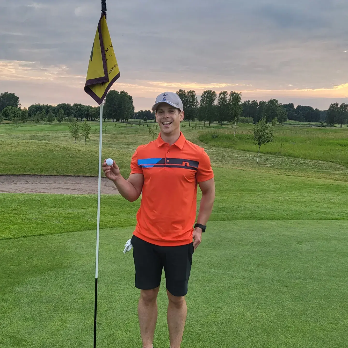 Mand i orange polo og kasket holder en golfbold ved siden af flagstangen på en golfbane med græs og træer i baggrunden.