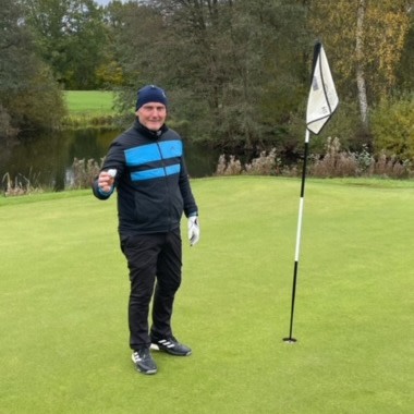 Man i golfkläder håller upp en golfboll och ler bredvid ett hål med flagga på en golfbana omgiven av träd.