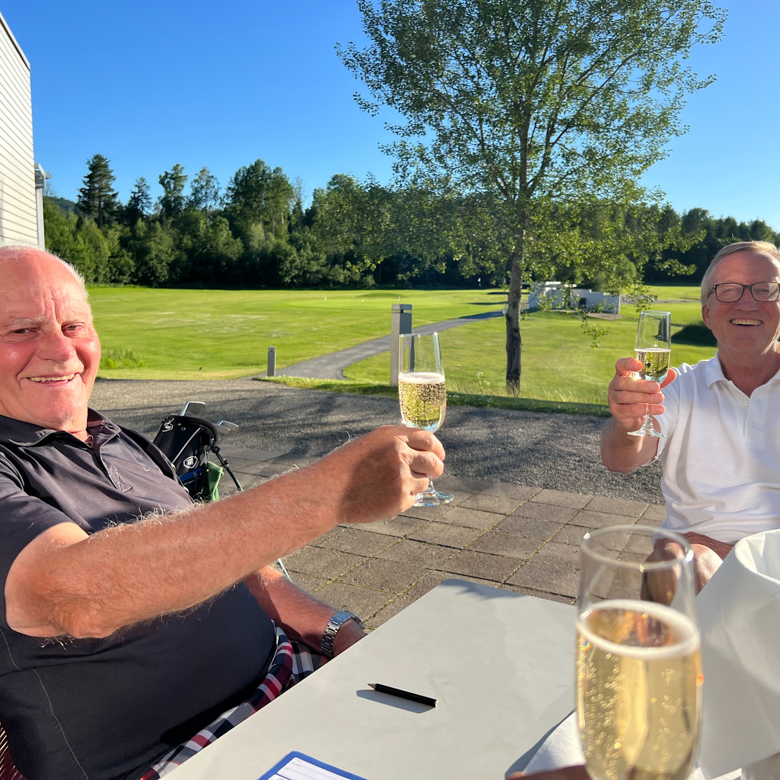 To ældre mænd skåler med champagneglas ved et bord udendørs på en solrig golfbane med grønne omgivelser i baggrunden.