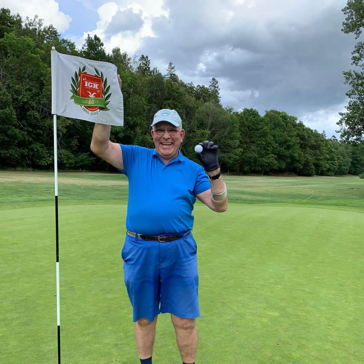 Ældre mand i blåt tøj holder en golfbold og et flag op på greenen, smilende, med skov i baggrunden.