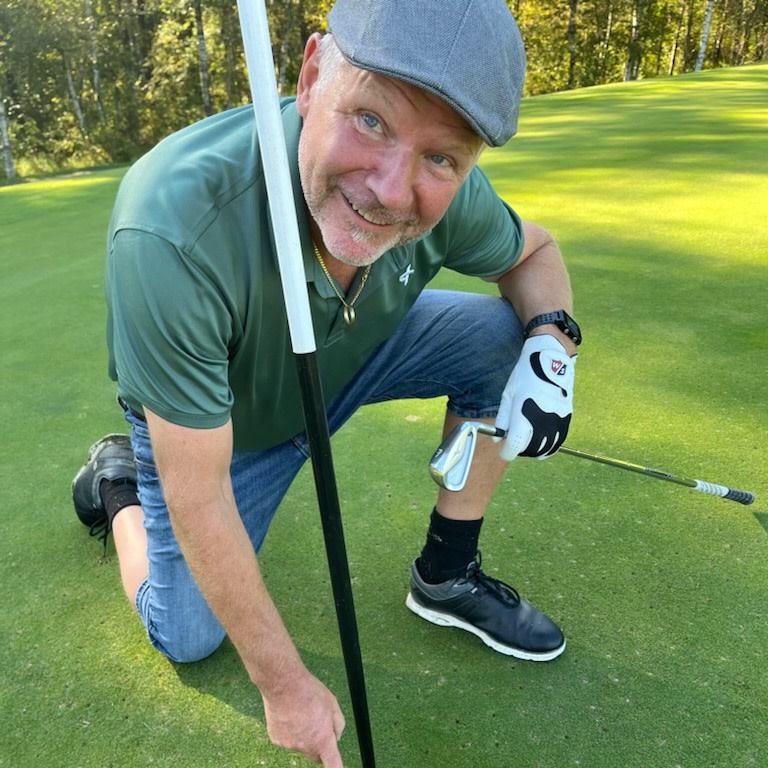 Man i keps och golfhandske knäböjer på en golfgreen, håller i flaggstången och pekar mot hålet med ett leende.