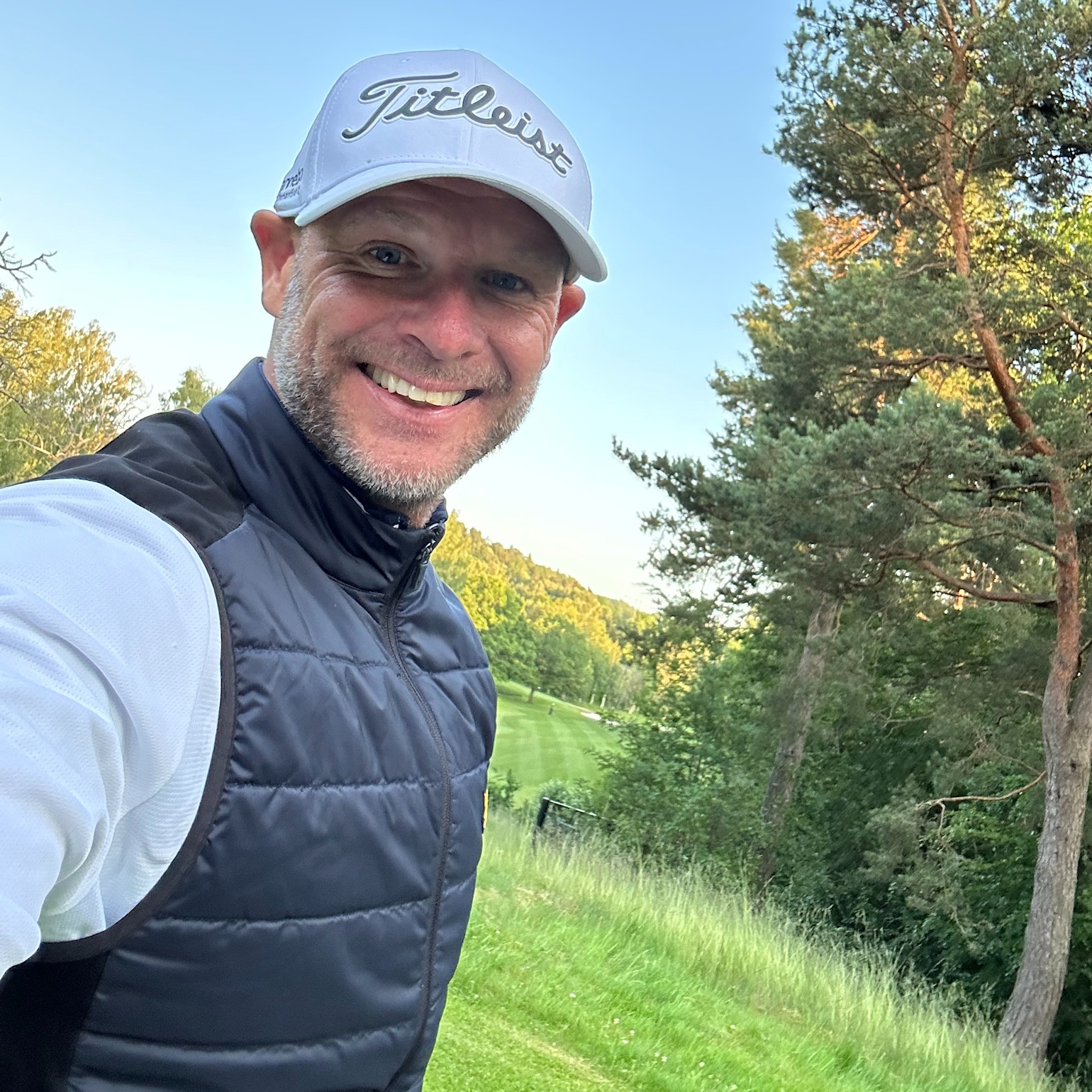 Leende man med keps med texten "Titleist" tar en selfie utomhus på en grön golfbana omgiven av träd.