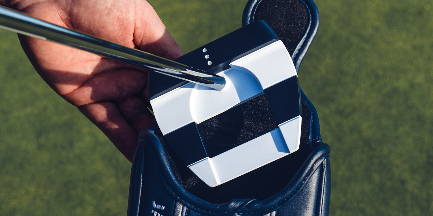En hand håller ett modernt golfputterhuvud med svart och vitt mönster ovanför ett blått fodral på en golfbana.