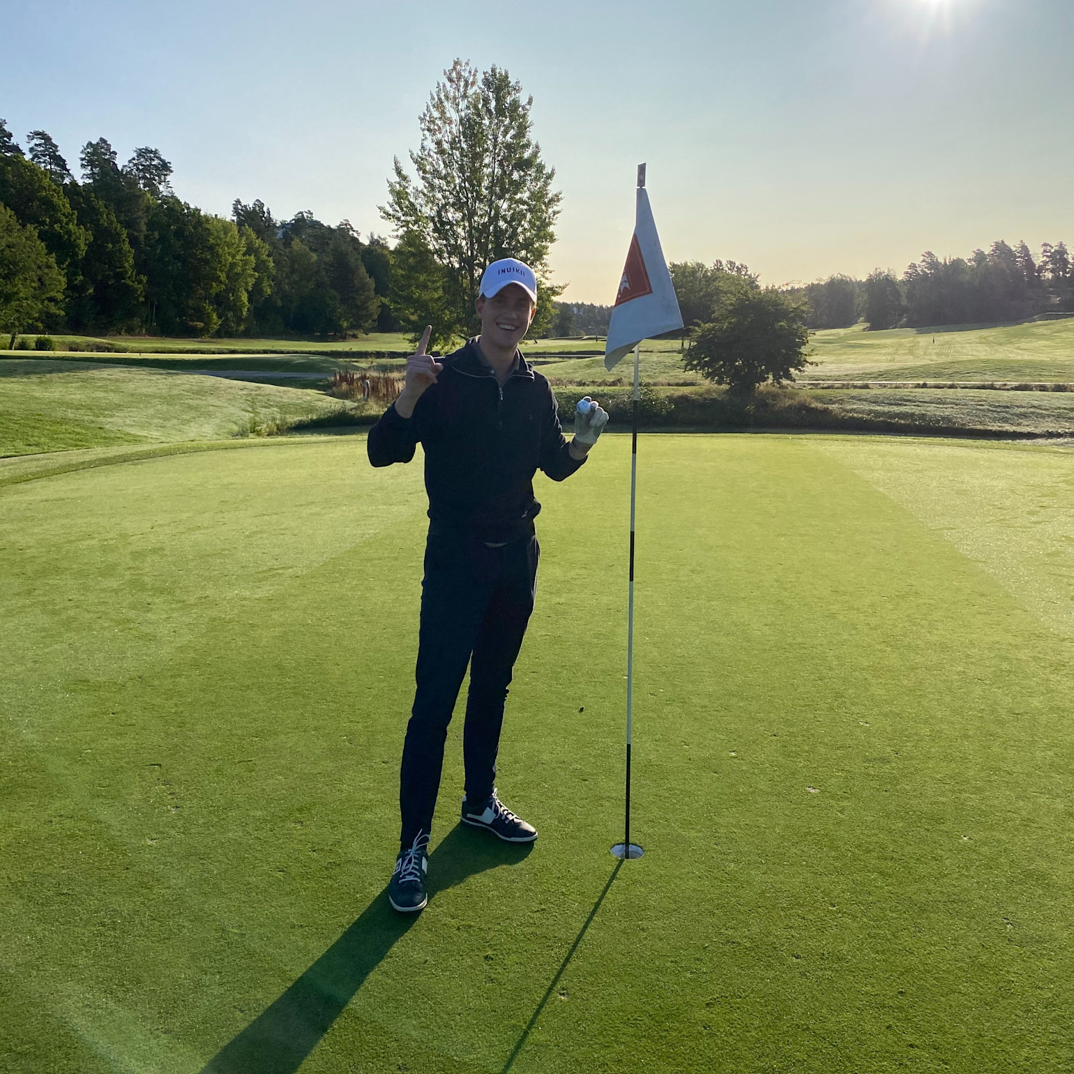 En person står på en golfgreen ved siden af flagstangen, holder en golfbold op og viser én finger, solrig morgen på golfbanen.