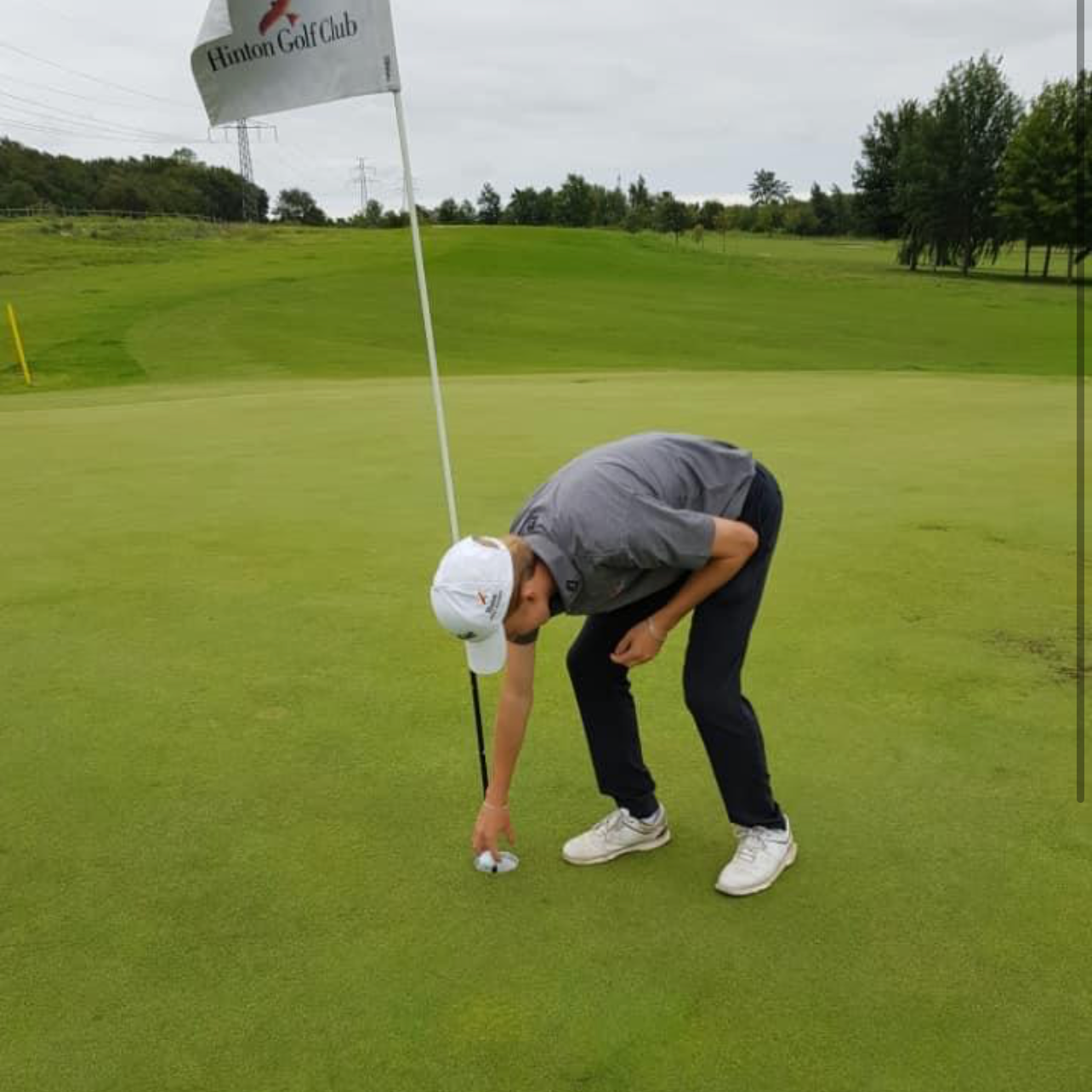 En golfspiller bøjer sig ned for at samle en golfbold op ved hullet på en grøn golfbane under en overskyet himmel.