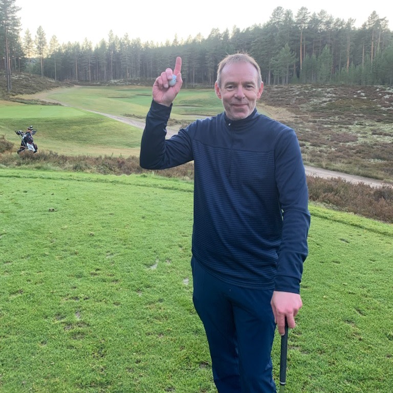 Mand iført mørkeblåt golf­tøj holder én finger og en golfbold op, står på en golfbane med skov i baggrunden.