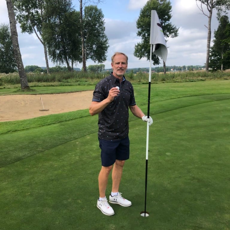 Man holder en golfbold og står ved siden af flagstangen på en green, med en sandbunker og træer i baggrunden.