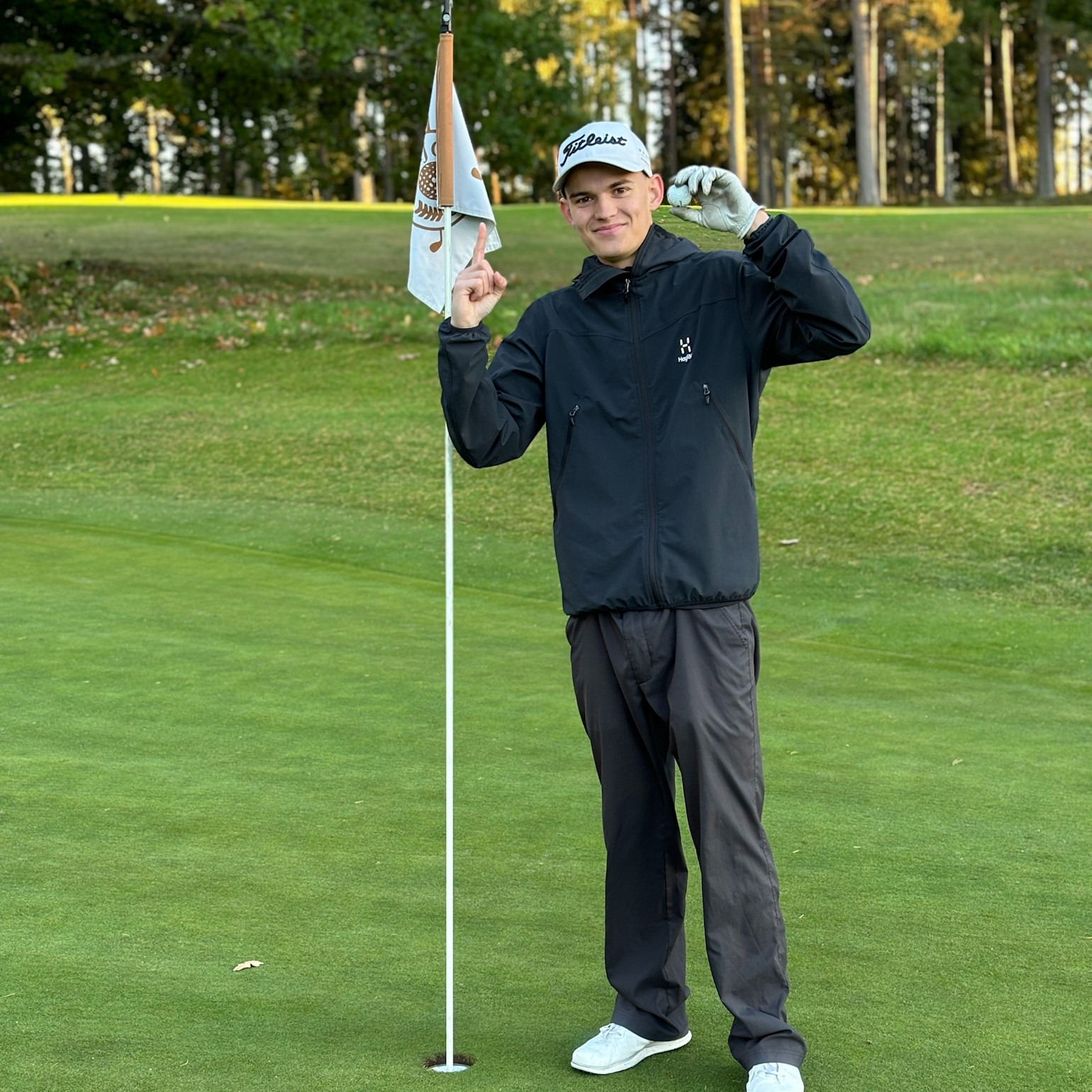 Ung man i golfkläder står bredvid ett hålflagg på en golfbana, håller upp en golfboll och pekar uppåt med ett finger.