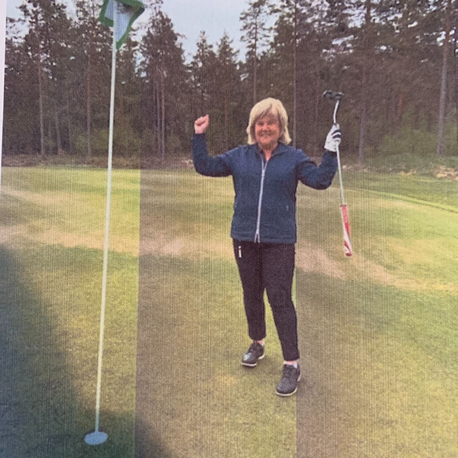 Kvinde fejrer med armene i vejret og holder en golfkølle ved siden af et golfhul på en grøn golfbane med skov i baggrunden.