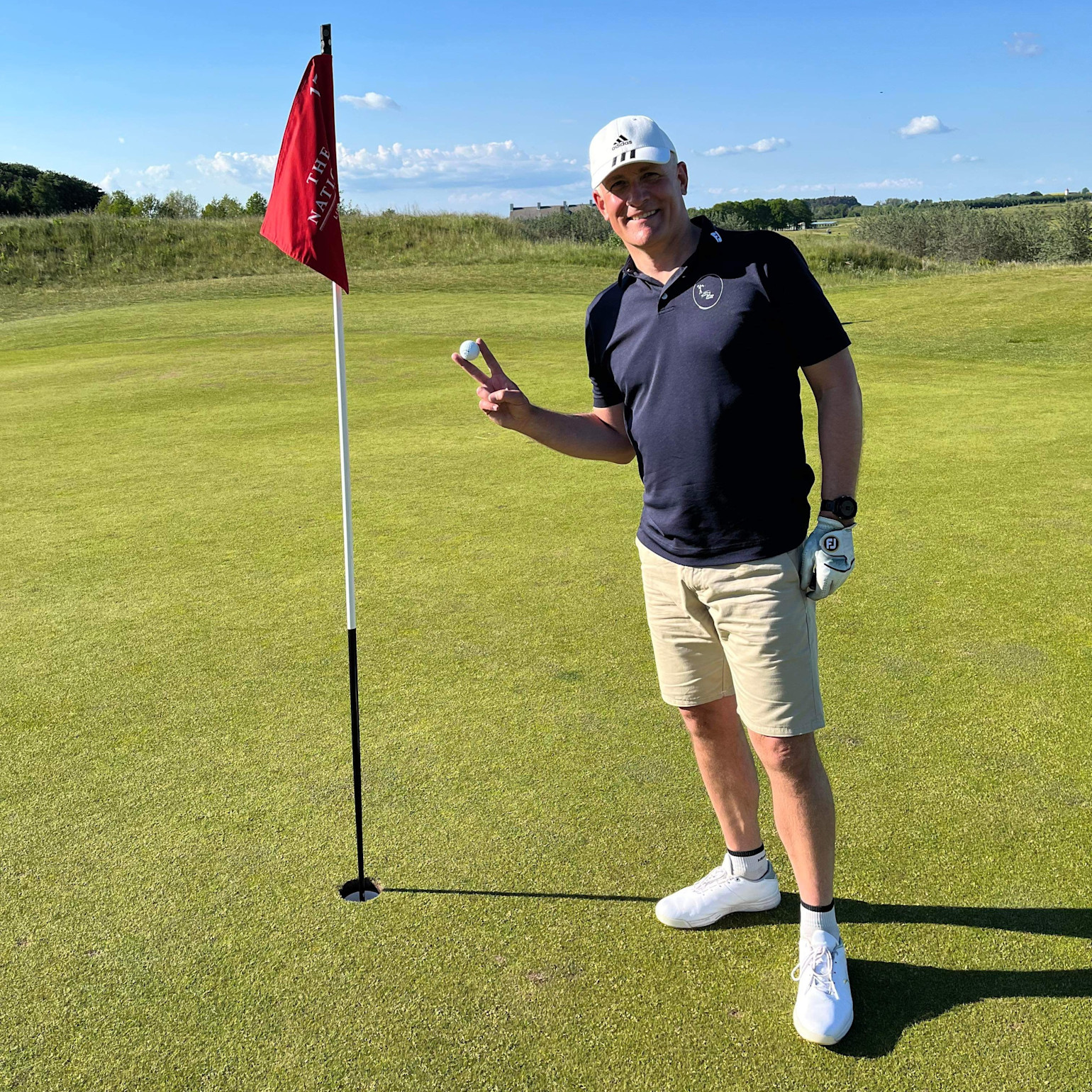 En mand i golfbeklædning holder en golfbold op og laver sejrtegn ved siden af et hul med rød flag på en golfbane i solskin.