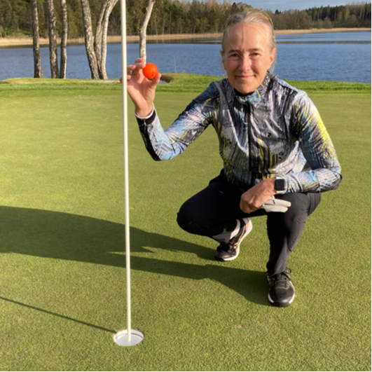 Personen knæler ved siden af et golfhul og holder en orange golfbold op på greenen, med en sø og træer i baggrunden.