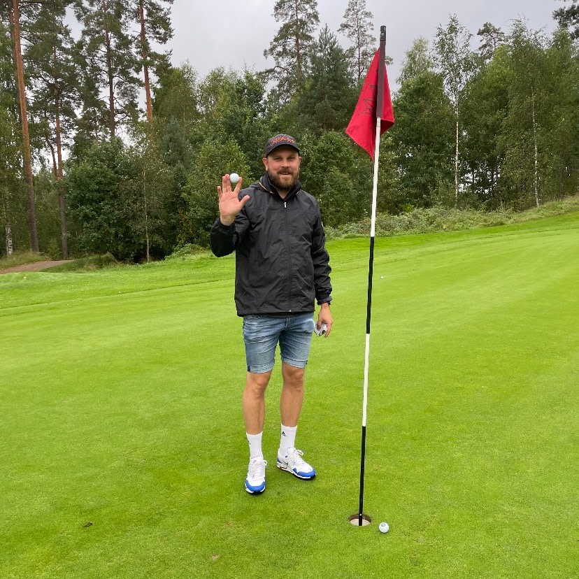 Mand i sort jakke og jeansshorts holder en golfbold op ved siden af et golfhul med flag på en grøn golfbane omgivet af træer.