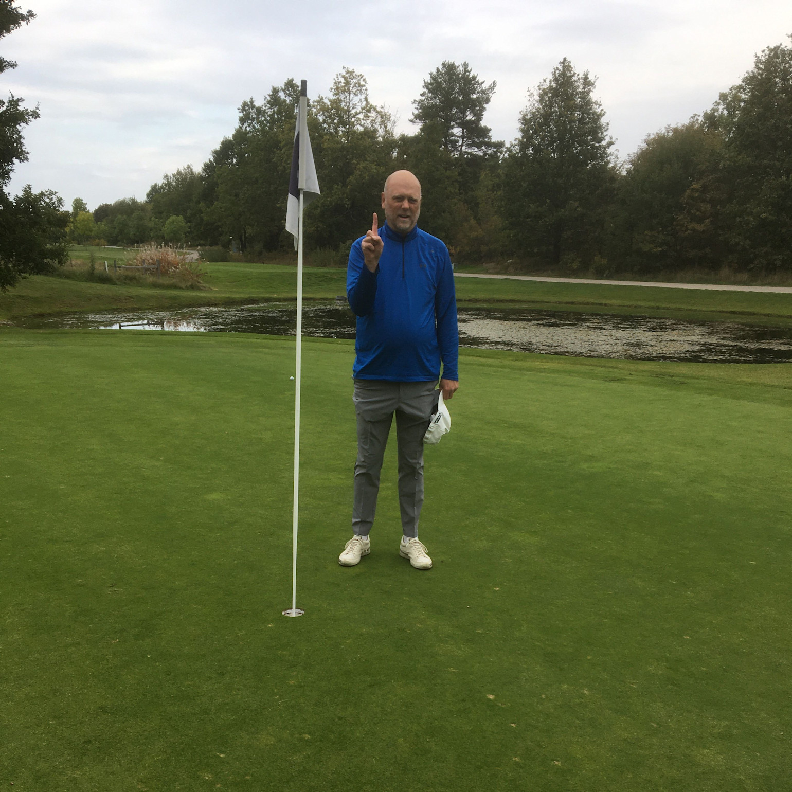 Mand i blå jakke står ved siden af et golf-flag på greenen og holder én finger op, med en sø og træer i baggrunden.