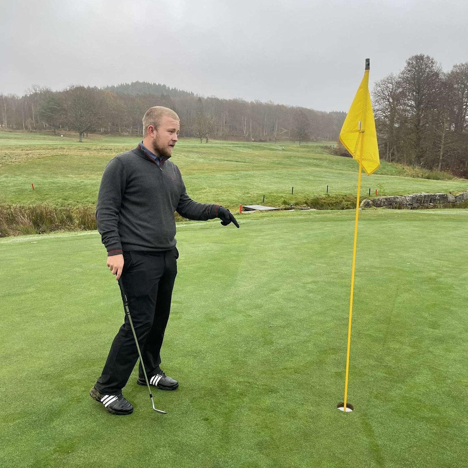 Man med golfklubba pekar mot golfhål bredvid gul flagga på green, omgiven av gräs och träd i bakgrunden.