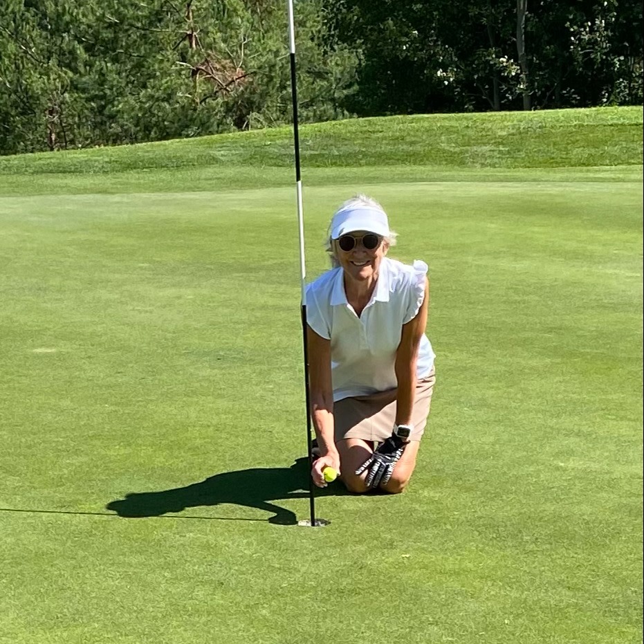 Kvinde i golfbeklædning knæler ved et golfhul og holder en golfbold på en solrig green med træer i baggrunden.