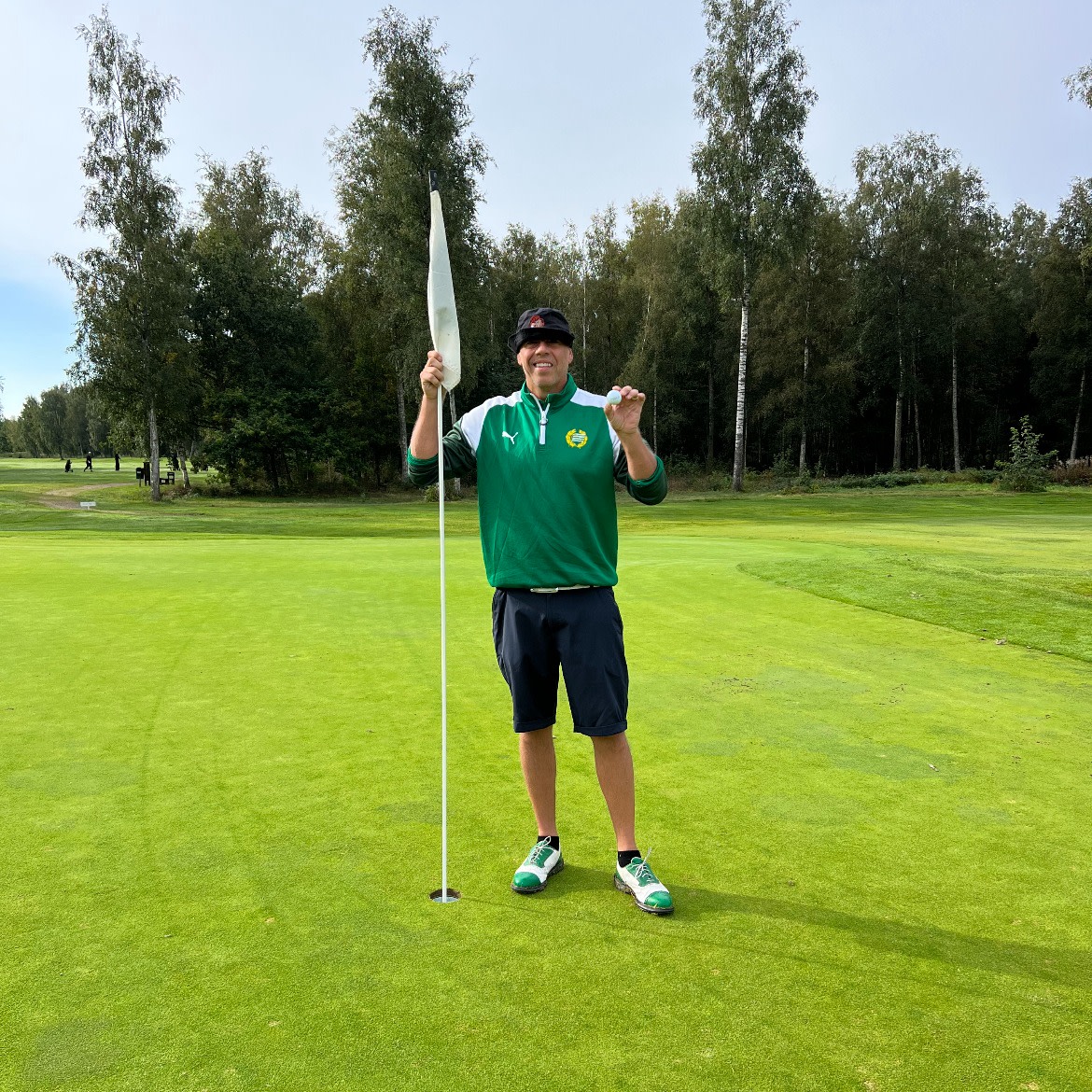 Man i grön tröja och keps håller en golfboll och flaggstång på en golfgreen, omgiven av träd och gräs.