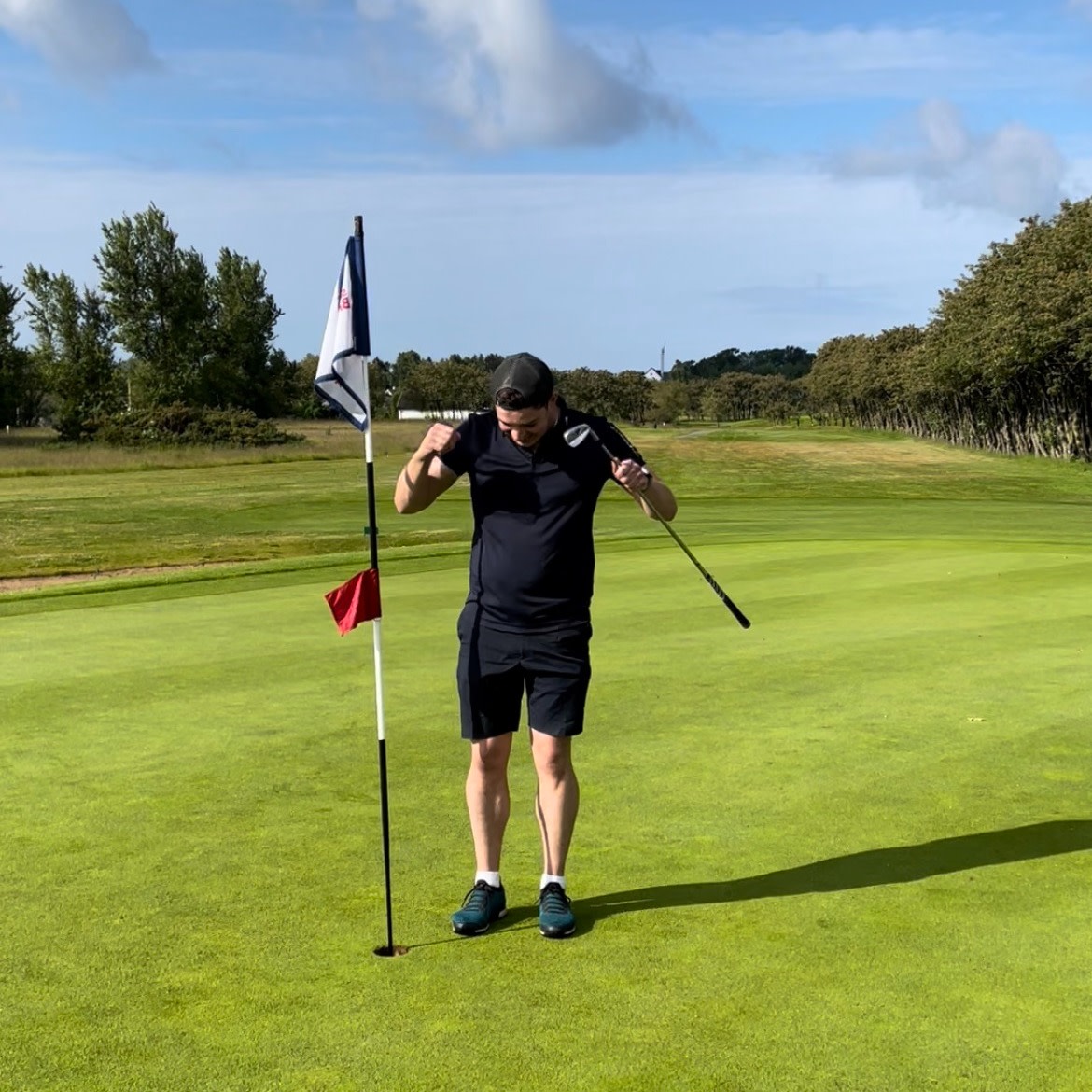 En mand i sorte golfklæder står ved en flagstang på en golfgreen og holder en golfkølle over skuldrene på en solrig dag.