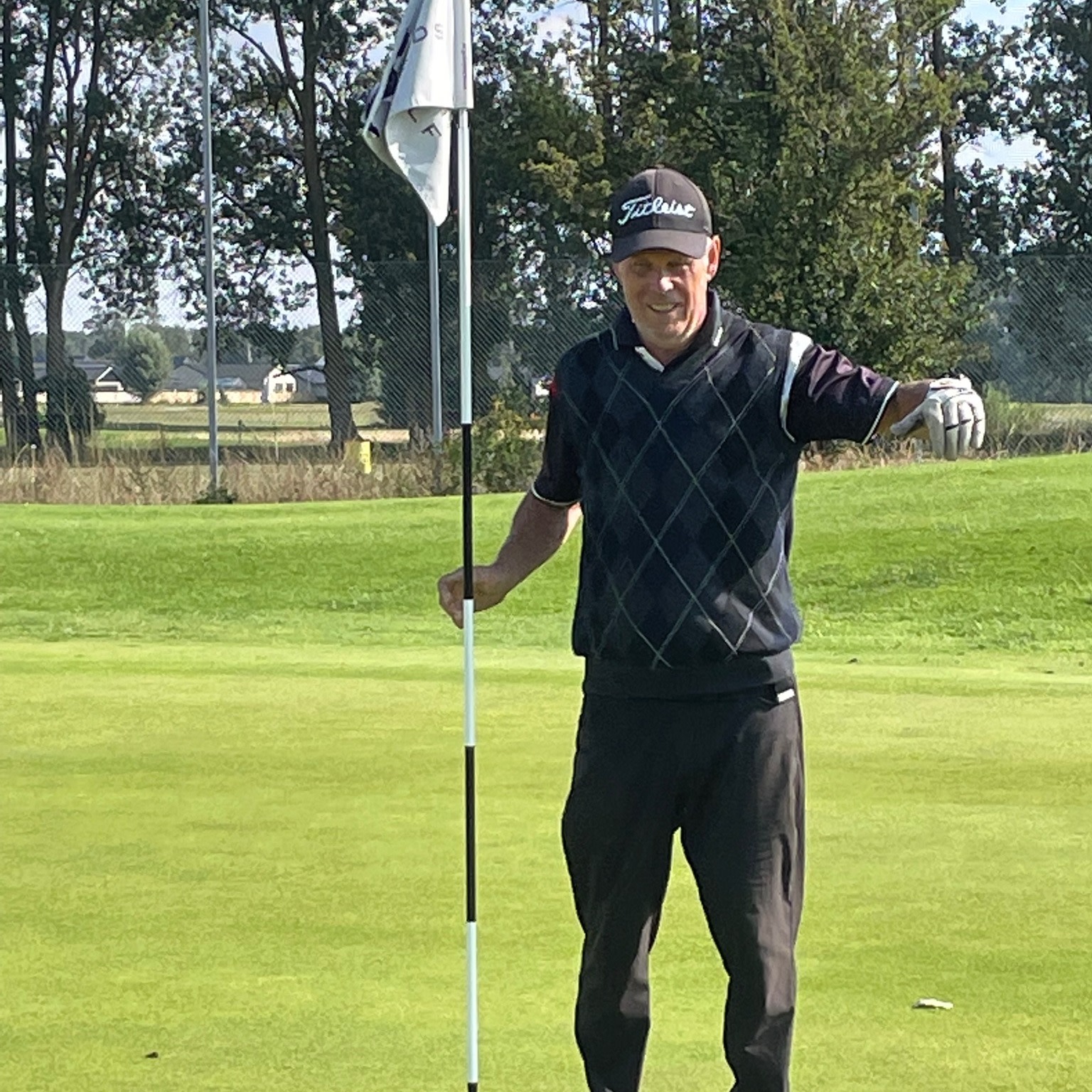 En mand iført golfbeklædning holder i flagstangen på en golfgreen, med træer og marker i baggrunden.