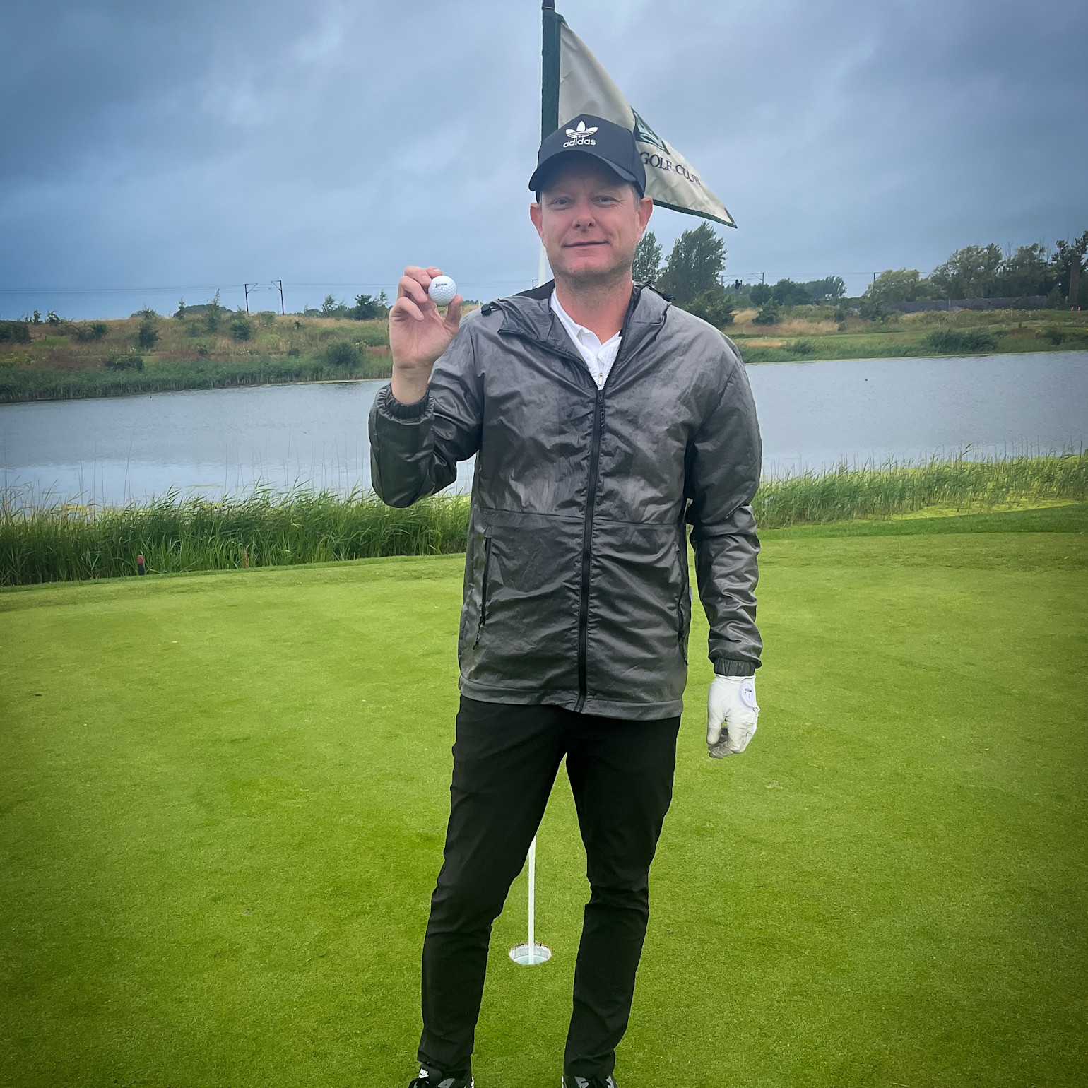 Man i golfkläder håller upp en golfboll på en green bredvid ett hål, med flagga och sjö i bakgrunden.