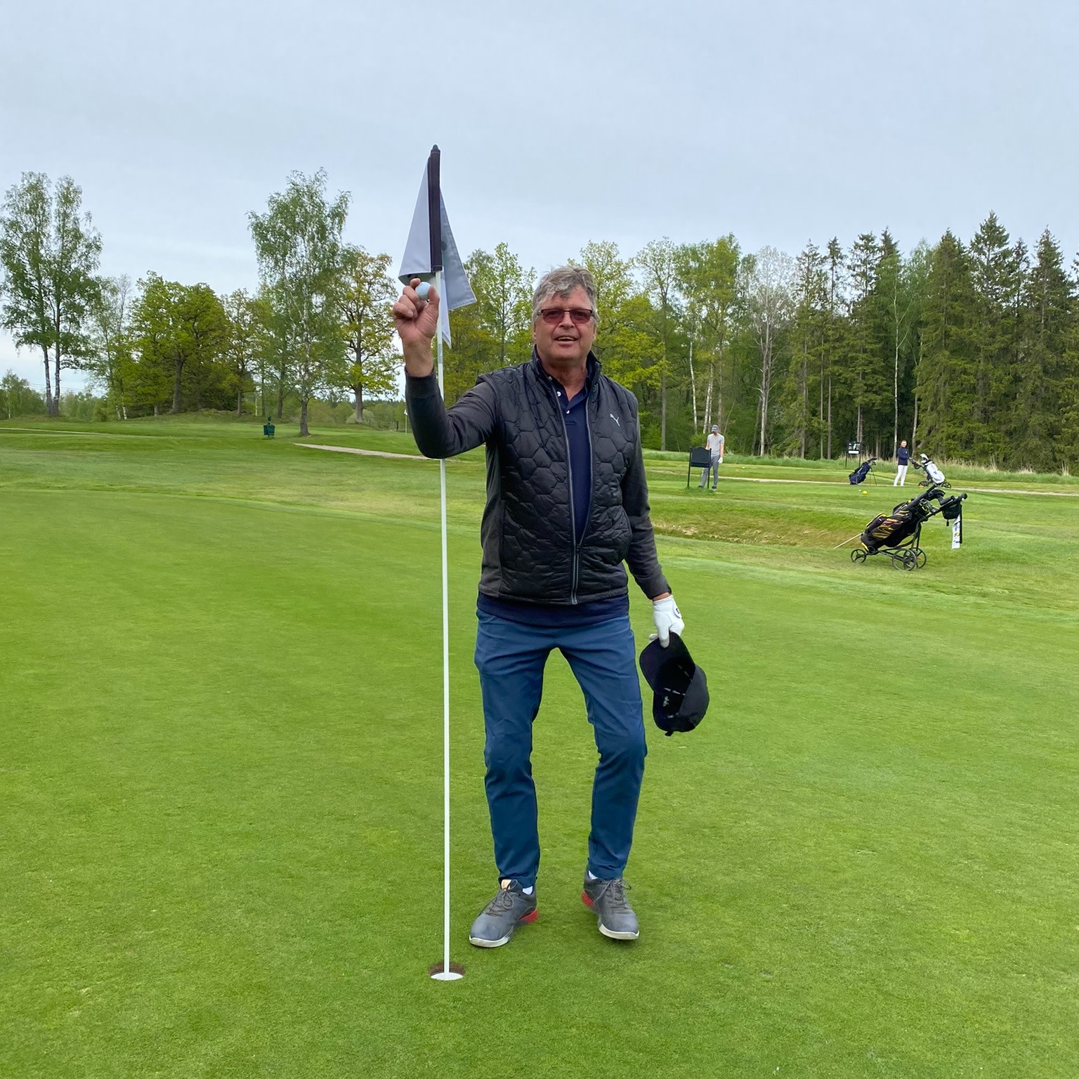 Man holder en golfbold ved flagstangen på en golfgreen, med en golfvogn og træer i baggrunden.